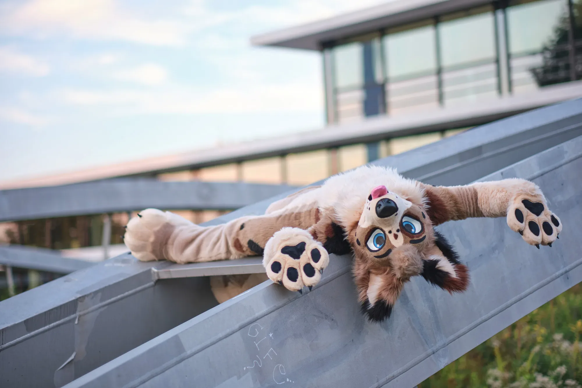A relaxed canine fursuiter lying on a stepped seating ledge outside a glass-fronted hotel at Eurofurence.
